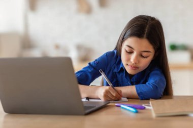 Çevrimiçi Okul. Liseli kız ders kitabına not alıyor laptopun yanında yazıyor evden ders çalışıyor, masa içinde oturuyor. Uzaktan Eğitim, Modern Öğrenim ve İnternet Teknolojisi