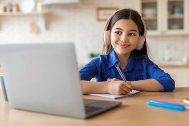 Başarılı bir E-öğrenme. Arapça Liseli Kız Evde Çevrimiçi Eğitimden Hoşlanıyor, Laptop kullanıyor ve sanal derste notlar alıyor, kapalı bir masada oturuyor. Uzaktan Okul Çalışması ve İnternet Teknolojisi