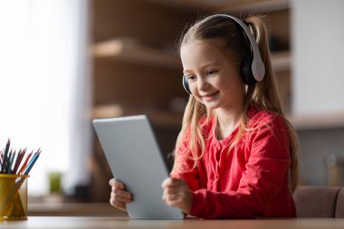 Küçük mutlu kız evde dijital tabletle dinleniyor, kablosuz kulaklık takan tatlı kız çocuğu online oyun oynuyor, modern cihazlarla video izleyen mutlu çocuk, kopyalama alanı.