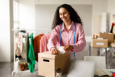 Moda sektörü. Neşeli Bayan Giysi Tasarımcısı karton kutuda moda giysileri paketliyor, mağazada çalışan müşteriler için sipariş hazırlıyor. Ticaret, Başarılı Girişimcilik