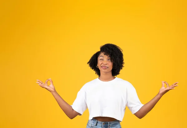 穿着白色T恤沉思的滑稽年轻的非洲卷曲女士笑着 用一只眼睛看着空旷的空间 孤立在黄色的工作室背景中 做广告和提供服务 — 图库照片