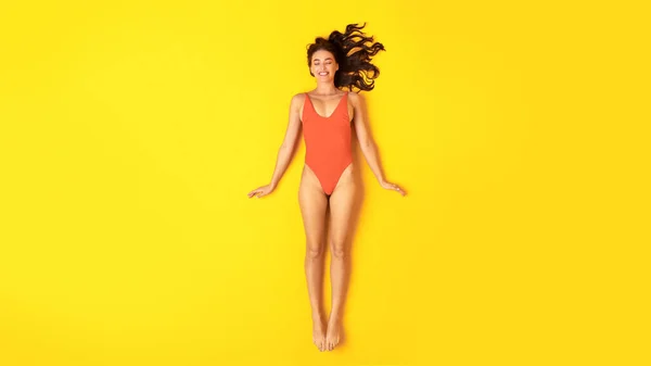 健康な夏の日光浴 オレンジ色のスリムな女1枚水着は目を閉じて横になり 黄色いスタジオの背景の上にあるプールで休暇を楽しんでいます 上を見るとパノラマ — ストック写真