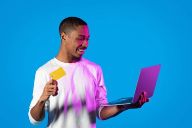 E-ticaret, perakende. Gülümseyen, sakallı, beyaz tişörtlü, banka kartı ve dizüstü bilgisayarlı, neon ışıkta mavi arka planda poz veren, internetten alışveriş yapan, fotokopi çeken yakışıklı adam.