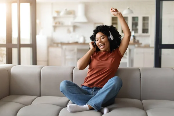 自宅でワイヤレスヘッドフォンで彼女のお気に入りの音楽を聞いて陽気な若い黒人女性 リビングルームでソファに座っている間に手で踊るケアフリーアフリカ系アメリカ人女性 スペースをコピー — ストック写真
