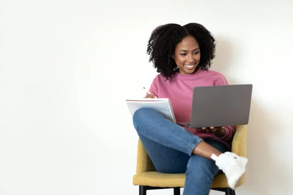 忙しい幸せな千年のアフリカ系アメリカ人のカジュアルな女性は アームチェアに座ってコンピュータを入力し 白い壁の背景にメモを取ります 仕事と勉強 ビデオレッスン ウェビナー 自宅での教育 — ストック写真