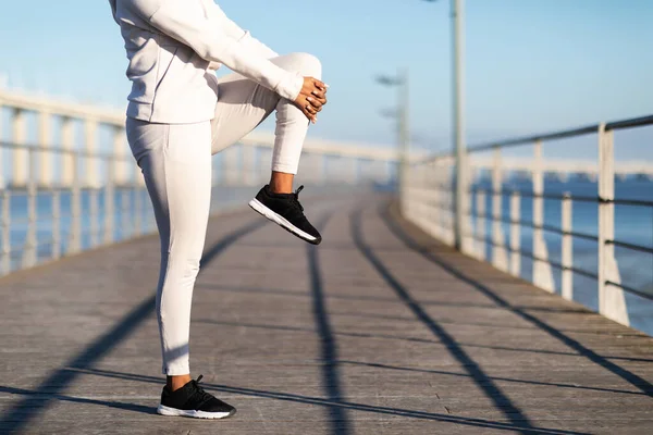 屋外で実行する前に認識できない黒の女性ストレッチ足の筋肉 スポーツウェアで若いアフリカ系アメリカ人女性は海の近くの木製の桟橋で作業 外でジョギングの準備クロップ — ストック写真