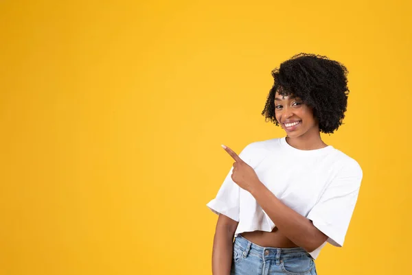 黄色のスタジオの背景に隔離された 空の空間で指を指して白いTシャツの中に千年アフリカ系アメリカ人の女性を笑顔 広告や提案 アドバイスやおすすめ — ストック写真