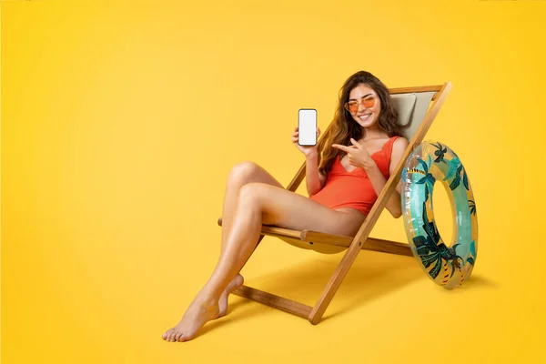 水着で陽気な若い混合レースの女性は サングラスのデッキチェアに座って 空白の画面を持つ携帯電話でポイント指 黄色の背景に隔離された休暇をお楽しみください 夏休みのためのアプリ Blog — ストック写真