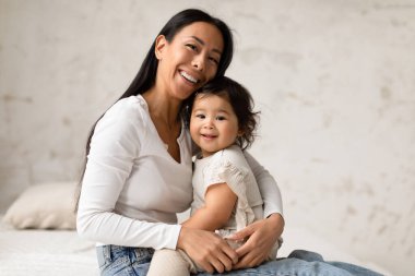 Aşk Bağları. Mutlu Asyalı anne ve bebek kızı kucaklaşıyorlar, kameraya gülümsüyorlar evde yatakta birlikte oturuyorlar. Ebeveynliğin ve Bebekliğin Neşeli Anları