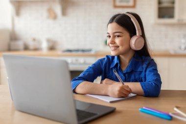 E-öğrenme. Kulaklıklı, bilgisayarlı, gülümseyen liseli kız. Evdeki masasında oturan uzaktan kumandalı video dersleri alıyor. Liseli Kız Uzaktan Ödev Yapmayı Çevrimiçi Öğreniyor