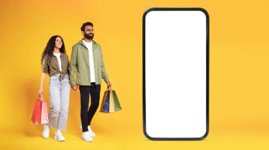 Moda Mağazası Başvurusu. Alışveriş torbası pozu veren, boş ekranlı akıllı telefonun yanında elini sarı stüdyoda tutan mutlu bir Arap çift. Mobil Ticaret. Panorama