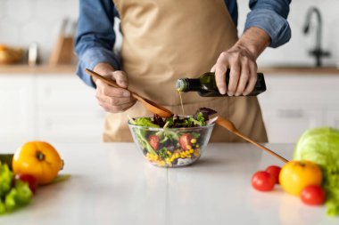 Sağlıklı gıda konsepti. Mutfakta yemek pişirirken sebze salatası tatlandıran olgun bir adam, önlüklü tanınmayan bir erkek aşçı evde vejetaryen yemeği hazırlıyor, zeytinyağı ekliyor, kırpılmış