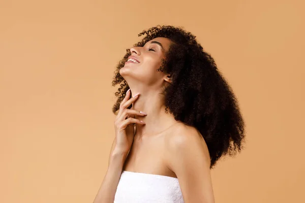 Joyful happy black lady wrapped in towel touching her soft flawless skin on neck, woman enjoying result of spa beauty treatments, posing on beige studio background, copy space