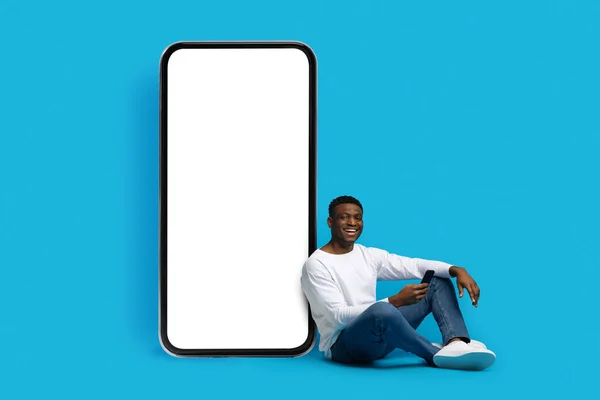 stock image Mobile app. Relaxed cheerful handsome young african american man in casual outfit sitting by huge cell phone with white empty screen over blue studio backgound, using smartphone, copy space, mockup