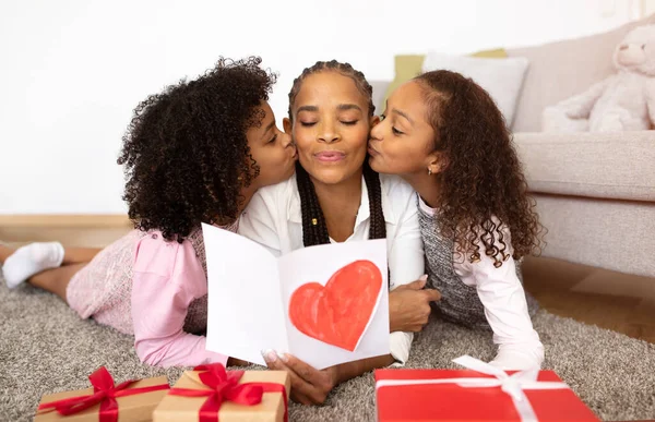 Moms Special Day. Excited African American Daughters Surprising Their Mother With a Crafted Card and Gifts On Her Birthday, Kissing Mommy On Cheeks Posing In Cozy Living Room At Home