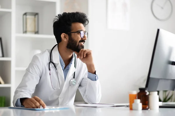 英俊的 友善的中东年轻医生 戴着眼镜坐在办公桌前 在现代化诊所的内部电脑上工作 与病人进行视频通话 并在网上预约 — 图库照片
