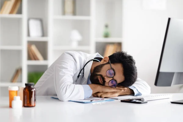疲惫不堪的年轻中东医生穿着医疗外套和眼镜躺在电脑前的桌子上 睡在现代诊所的工作场所 保健工作者和精疲力竭 — 图库照片