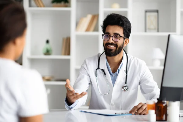 Young arab man doctor consulting lady patient, filling form at consultation. Handsome bearded family doctor man wearing white coat talking to woman while appointment visit in modern clinic