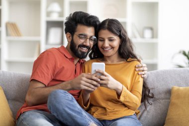 Genç gülümseyen Hintli çift cep telefonu kullanıyor ve evde kucaklaşıyorlar, mutlu doğu eşleri modern akıllı telefonlarıyla koltukta dinleniyor, yeni uygulamalara bakıyor ya da internetten alışveriş yapıyor, Space 'i kopyalıyor.