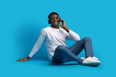 Neşeli, mutlu, yakışıklı, Afro-Amerikalı, sıradan bir şekilde oturmuş mavi stüdyo arka planında müzik dinliyor, kablosuz kulaklık kullanıyor ve gülümsüyor.