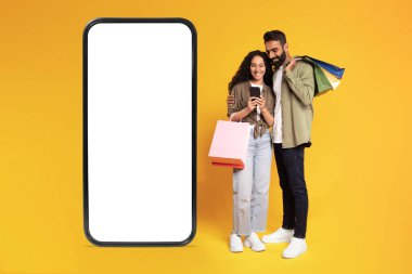 Alışveriş Uygulaması. Sarı Stüdyo Arkaplanı üzerinde boş ekranı olan, cep telefonu satın alma giysileri kullanan neşeli genç bir Arap çift, alışveriş torbası pozu veriyor. Model
