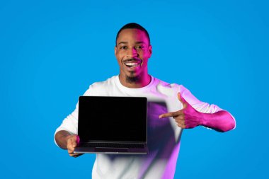 Heyecanlı mutlu Afro-Amerikan genç adam siyah beyaz ekranlı bilgisayar defterini işaret ediyor ve gülümsüyor, güzel bir web sitesi tavsiye ediyor, sohbet robotu, mavi arka plan, maket, kopyalama alanı