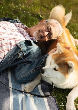 Gözlüklü, sakallı, neşeli, beyaz bir adam köpekle ekoseye uzanıyor, dinleniyor, rahatlıyor, parkta, dışarıda piknik yapıyor. Hayvan, aşk ve eğlenceyle hafta sonu yürüyüşü, aktif yaşam tarzı.
