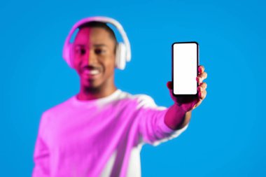 Beyaz beyaz ekranlı akıllı telefon Afro-Amerikan erkek eli, kablosuz kulaklık kullanan neşeli genç siyah adam telefon gösteriyor, güzel bir müzik uygulaması öneriyor, mavi arka plan, model.