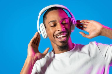 Duygusal genç siyahi adamın yeni kulaklıklarının keyfini çıkarması, kapalı gözlerle müzik dinlemesi, kulaklığa dokunması ve mavi arka planda neon ışığı altında şarkı söylemesi, boşluğu kopyalaması.