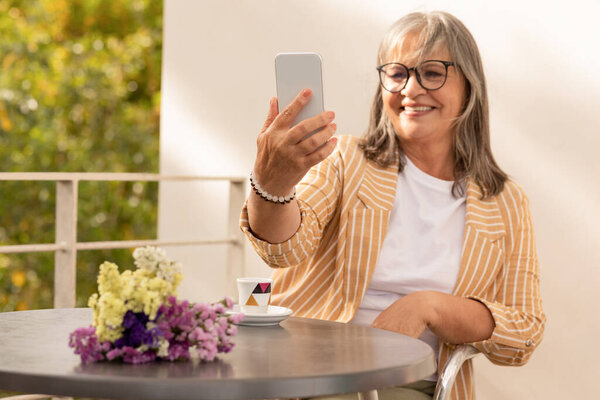 Cheerful european mature woman sit in cafe with flowers, drink coffee, take selfie on smartphone, enjoy social networks, outdoor. Chat app and video call remote at cafe at summer