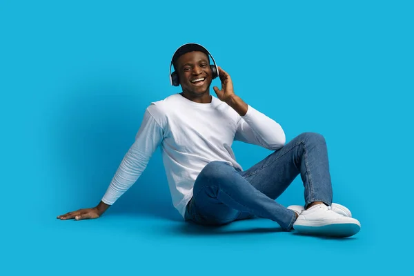 喜びに満ちたハンサムな若いアフリカ系アメリカ人男性で 床に座って 青いスタジオの背景の音楽を聞いて ワイヤレスヘッドフォンと笑顔を使用して コピースペース — ストック写真
