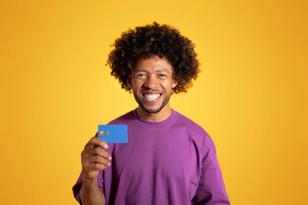 Cheerful black adult curly man in purple t-shirt shows credit card, isolated on orange background, studio. Shopping recommendation, finance cashback and profit, ad and offer