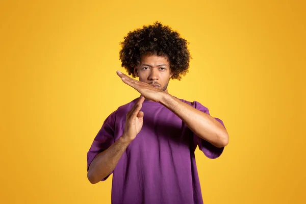 Serious black adult curly man in purple t-shirt show crossed hands, stop gesture, time out sign, isolated on orange background, studio. Break in quarrel, no, advertising and offer