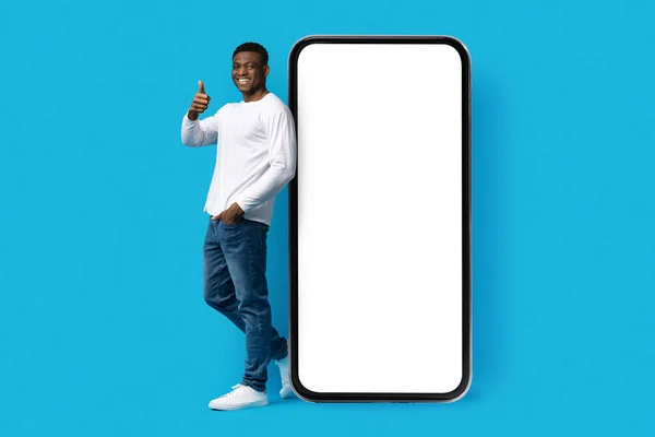 不错的移动应用程序 在线提供 积极英俊的年轻非洲裔美国人 站在一个巨大的手机旁边 屏幕空荡荡 露出大拇指 在蓝色的工作室背景下与世隔绝 — 图库照片