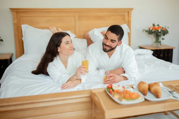 ホテルルームサービス リラックスしたカップルはベッドでロマンチックな朝食を楽しみます 新鮮なオレンジジュースを飲むベッドルームのインテリアでバスローブを身に着けて横たわっています おいしい食べ物とトレイ近く 新婚旅行の休暇 — ストック写真