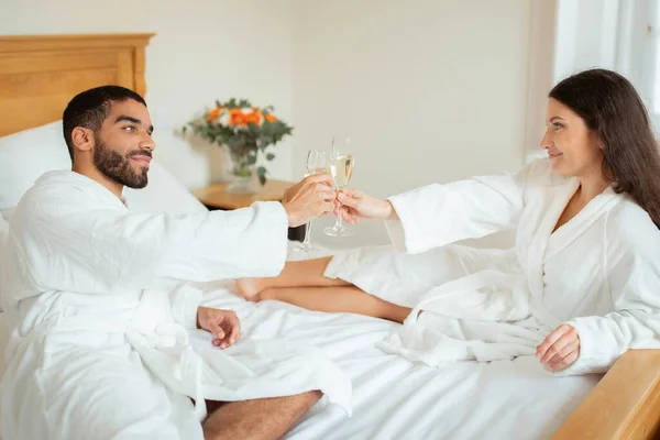 ロマンチックな新婚旅行 新婚夫婦は親密な瞬間を共有します シャンパングラスをクリック ホテルのベッドルームのインテリアで一緒にスパークリングワインをお楽しみください 休暇のレジャーとリラックス — ストック写真