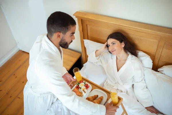 新婚旅行休暇レジャー お二人はベッドで朝食をとり 夫は妻に食べ物を渡し ホテルの部屋の快適さを楽しみ 柔らかい白いバスローブを着て — ストック写真