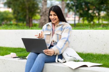 Gülümseyen İspanyol öğrenci kız dışarıda dizüstü bilgisayarla video görüşmesi yapıyor, mutlu kadın telekonferans için bilgisayarı kullanıyor, parkta oturuyor, online iletişimin keyfini çıkarıyor.