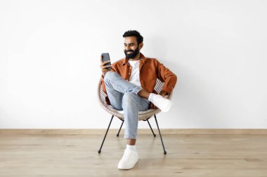 Hafta sonu, dijital eğlence. Akıllı telefon kullanan mutlu Hintli adam evde online eğlencenin tadını çıkarıyor. Cep telefonuyla mesajlaşan, beyaz duvarın karşısında oturan genç bir adam.