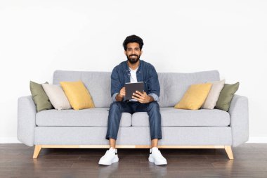 Çevrimiçi asistan, yapay zeka. Evdeki dijital tableti kullanarak gündelik kıyafetlerini giymiş neşeli, yakışıklı, genç doğu çocuğu. En yeni sohbet robotlarını, web sörflerini, kopyalama alanlarını kontrol ediyor.