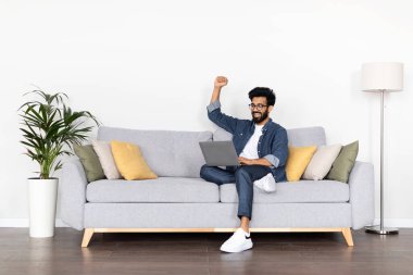 Online ticaret, kumar. Duygusal açıdan mutlu, orta doğulu, sıradan kıyafetler giyen ve gözlüklü, dizüstü bilgisayarlı koltukta oturan, el kol hareketi yapan, bağıran, ev içi, fotokopi çeken genç adam.