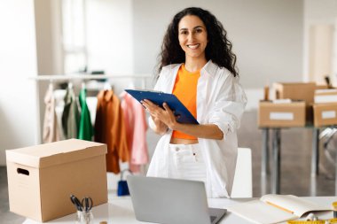 Başarılı Moda İşkadını 'yım. Giysi mağazasında yönetici olarak çalışan gülümseyen bir kadın elinde bir pano, elinde kameraya gülümseyen, elinde dizüstü bilgisayar ve karton kutuyla masanın yanında duran bir kadın. Perakende İş Teklifi