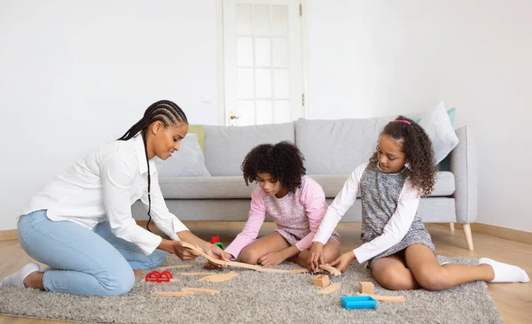 Happy African American Mom And Two Daughters Playing Together With Toy Railway Having Fun On Weekend, Sitting On Floor In Cozy Modern Living Room. Mommy Enjoying Pastime With Preteen Kids At Home