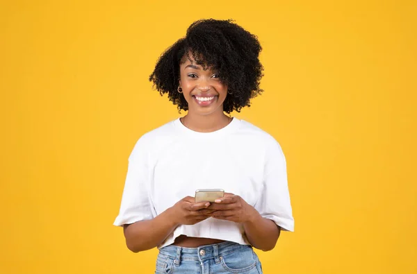 黄色のスタジオの背景に隔離されたスマートフォンで入力する白いTシャツの千年アフリカ系アメリカ人の巻き毛の女性を笑顔 広告とオファー ソーシャルメディアアプリとブログ ガジェットでのコミュニケーション — ストック写真