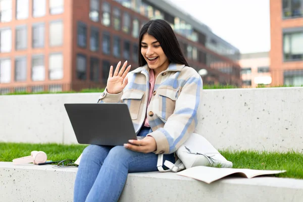 興奮した学生の女性は オンラインレッスン 教師とのビデオ通話 現代大学の公園に座っている間にノートパソコンに手を振って 距離研究とEラーニングの概念 — ストック写真