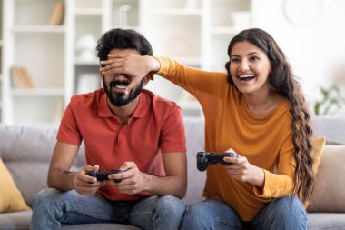 Neşeli Hintli çift evde eğleniyor, video oyunları oynuyorlar, kaygısız genç Doğu eşleri joystickleri kullanıyor ve oturma odasında otururken birbirleriyle yarışıyorlar.