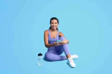 Spor giyimli, spor giyinen, su içen, telefonda yazan, mavi stüdyo arka planında izole bir şekilde oturan binlerce yıllık beyaz kadın. Spor, kilo kaybı ve vücut bakımı uygulaması
