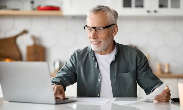 引退後の事業 キッチンのインテリアでラップトップで働く笑顔のシニア男性の肖像 デスクに座って リモートジョブのためのコンピュータを使用してハンサムな高齢ビジネスマンオンライン — ストック写真