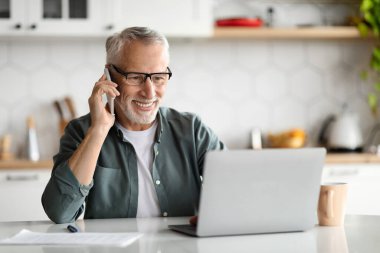 Evde dizüstü bilgisayarla çalışan ve cep telefonuyla konuşan yakışıklı, yaşlı ve yakışıklı bir beyefendi mutfak masasında oturuyor, uzaktan çalışmanın ve iş iletişiminin tadını çıkarıyor,