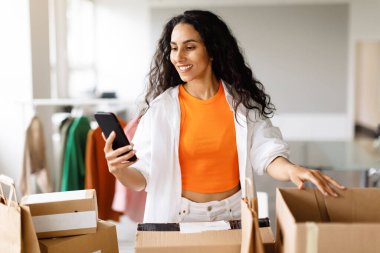 Ticaret için. Akıllı Telefon Uygulaması Karton Kutuları Kullanan Neşeli Arap Girişimci Kadın Moda Showroom 'da Giysi Siparişlerini Çevrimiçi Yönetmek. Başarılı Kıyafet Perakende Şirketi